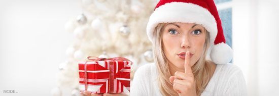 Woman in Santa hat holding a wrapped gift, finger on lips, Christmas tree background.