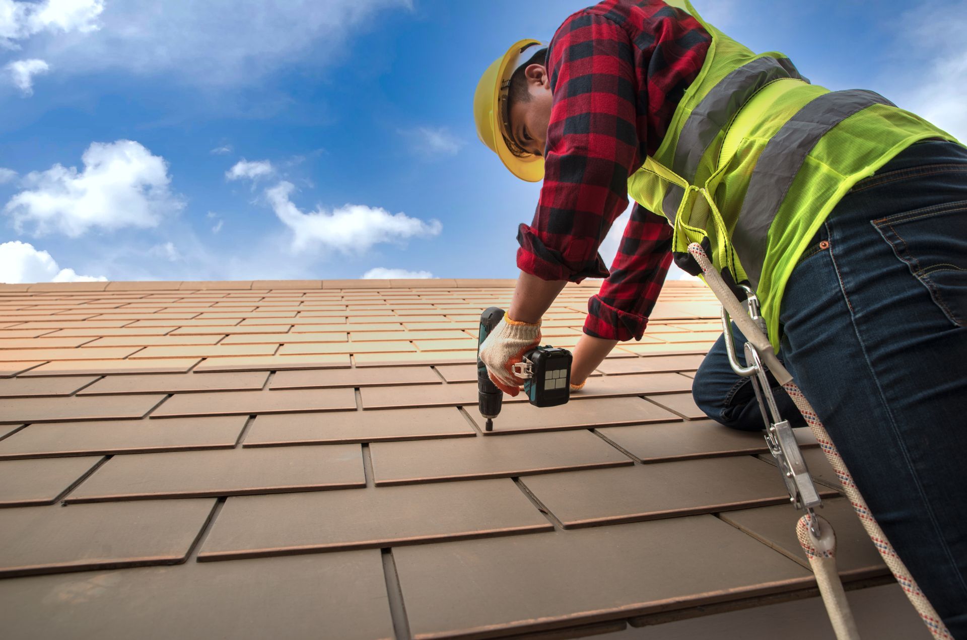 Worker using a power drill on a shingled roof under a blue sky