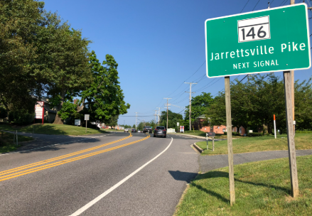 Road 146 sign for Jarrettsville Pike on a sunny suburban street with trees and a curving road