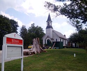 Small white church with steeple beside a Wilson sign and large tree stump on a grassy lawn