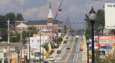Busy town street with cars, storefronts, church steeples, and an American flag on a cloudy day