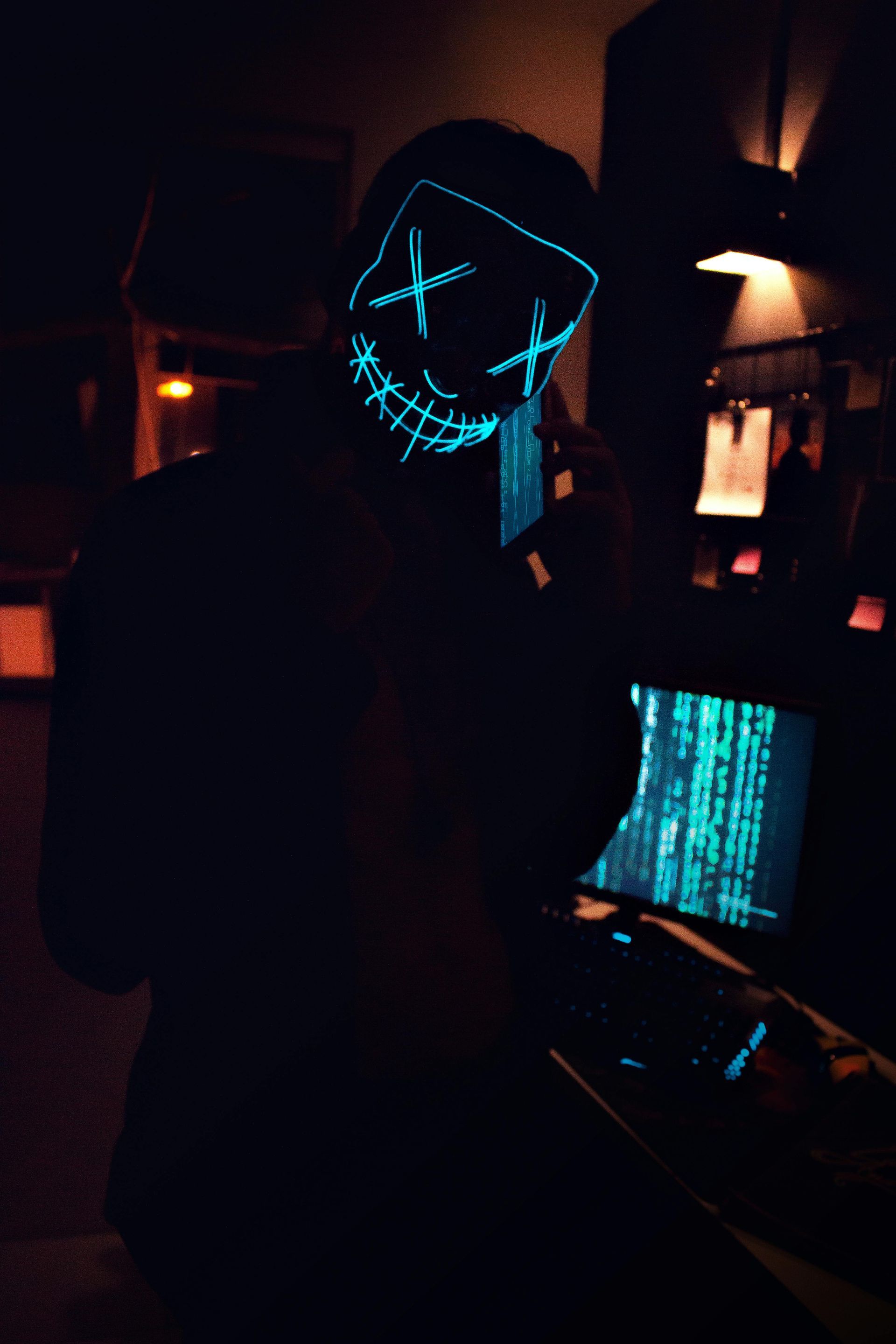 Hacker wearing an illuminated neon mask with crossed-out eyes, holding a smartphone in a dark room with computer screens displaying coding, symbolizing cybercrime, hacking, and digital security threats