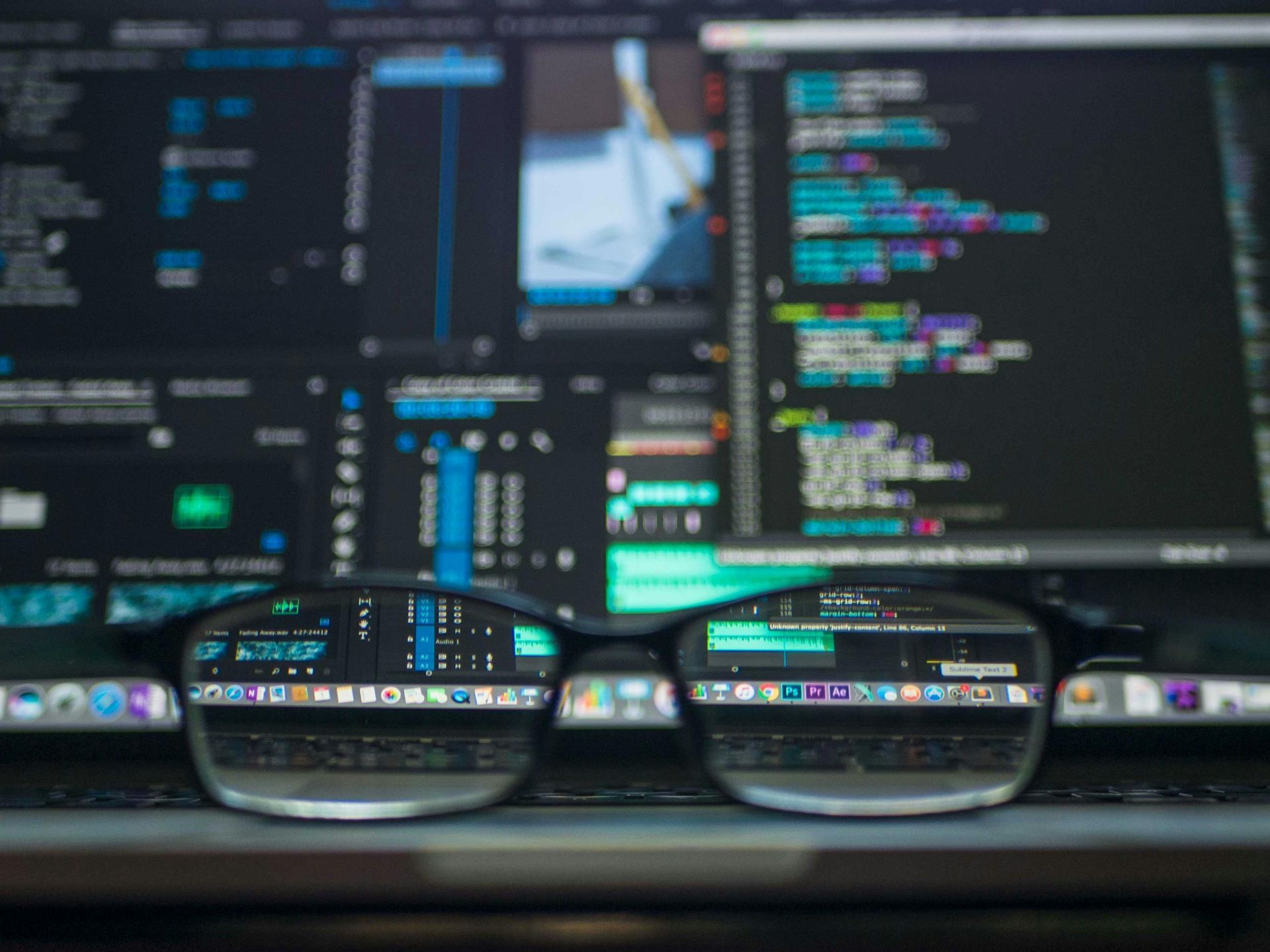 Eyeglasses resting on a laptop keyboard, reflecting colorful programming code and development tools on dual computer screens, symbolizing coding focus and digital work