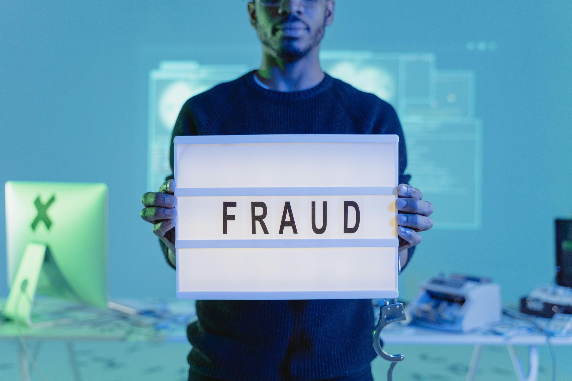 Man holding a lightbox sign displaying the word 'Fraud' with a handcuff on his wrist, standing in a tech-themed room with computer screens in the background, symbolizing online scams and cybersecurity risks.