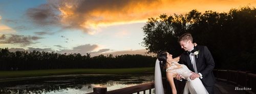 a bride and groom are posing for a picture on a bridge overlooking a lake at sunset .