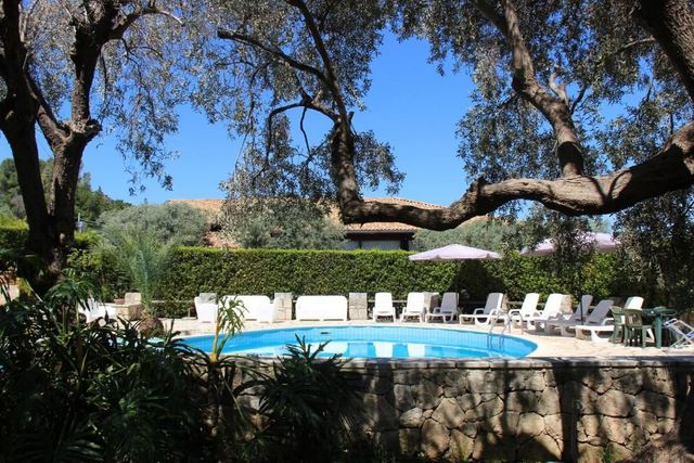 Piscina in giardino con lettini bianchi sotto alberi frondosi e un cielo azzurro.