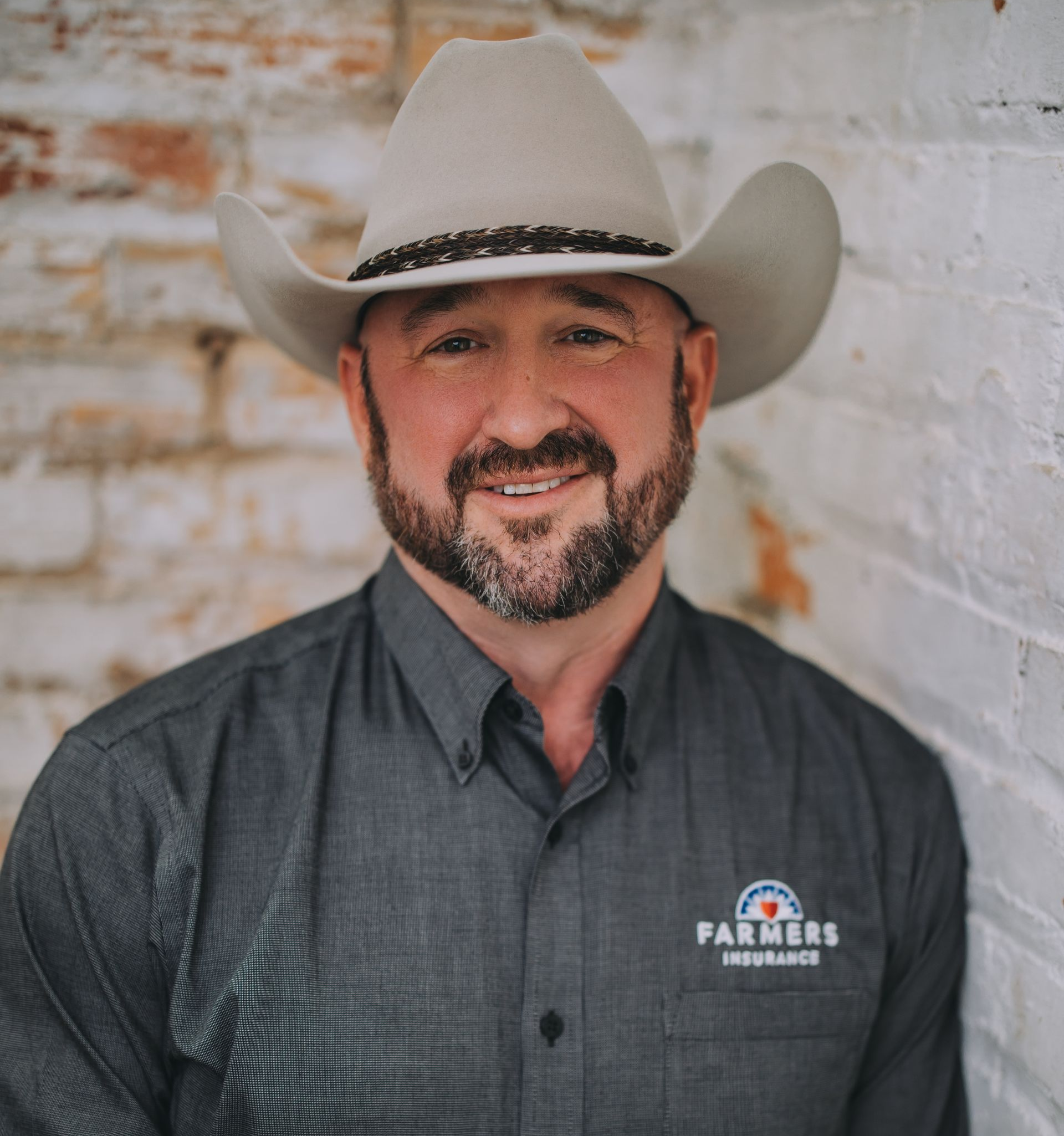 Man in cowboy hat, Farmers Insurance shirt, smiling by a brick wall.