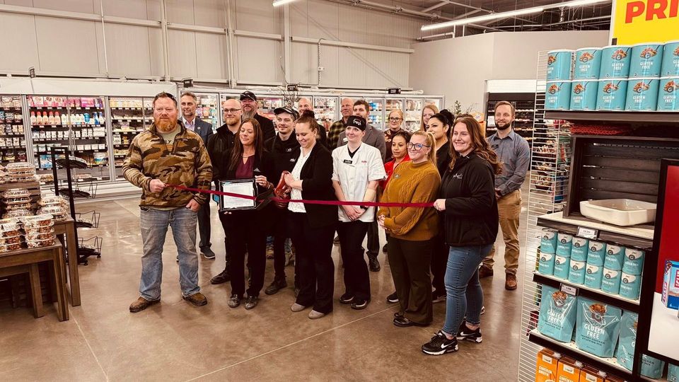 Group cutting a red ribbon at a store opening. Several people smile, shelves stocked with products visible.