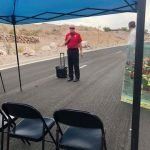 A man with a suitcase is standing on the side of a road under a blue tent.