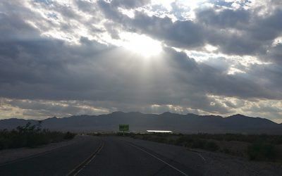 The sun is shining through the clouds over a desert road.