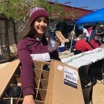 A woman is holding a cardboard box in front of a table.