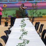 A long white table with green leaves on it is sitting in front of a colorful wall.