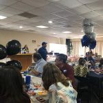 A group of people are sitting at tables in a room with balloons on the ceiling.