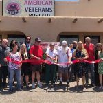 A group of people are standing in front of a building holding a red ribbon.
