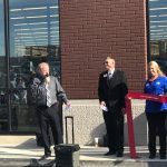 A group of people are standing in front of a brick building cutting a red ribbon.
