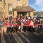 A large group of people are standing in front of a house holding a red ribbon.