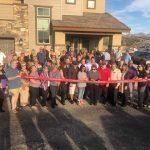 A large group of people are standing in front of a house holding a red ribbon.