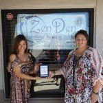 Two women are standing in front of a store holding a tablet.
