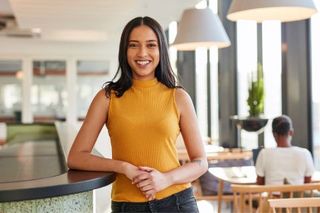 Woman in yellow top smiles, leans on counter in bright office space, person in background.