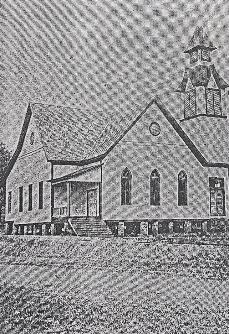 A black and white photo of a wooden church with a steeple on a grassy field.