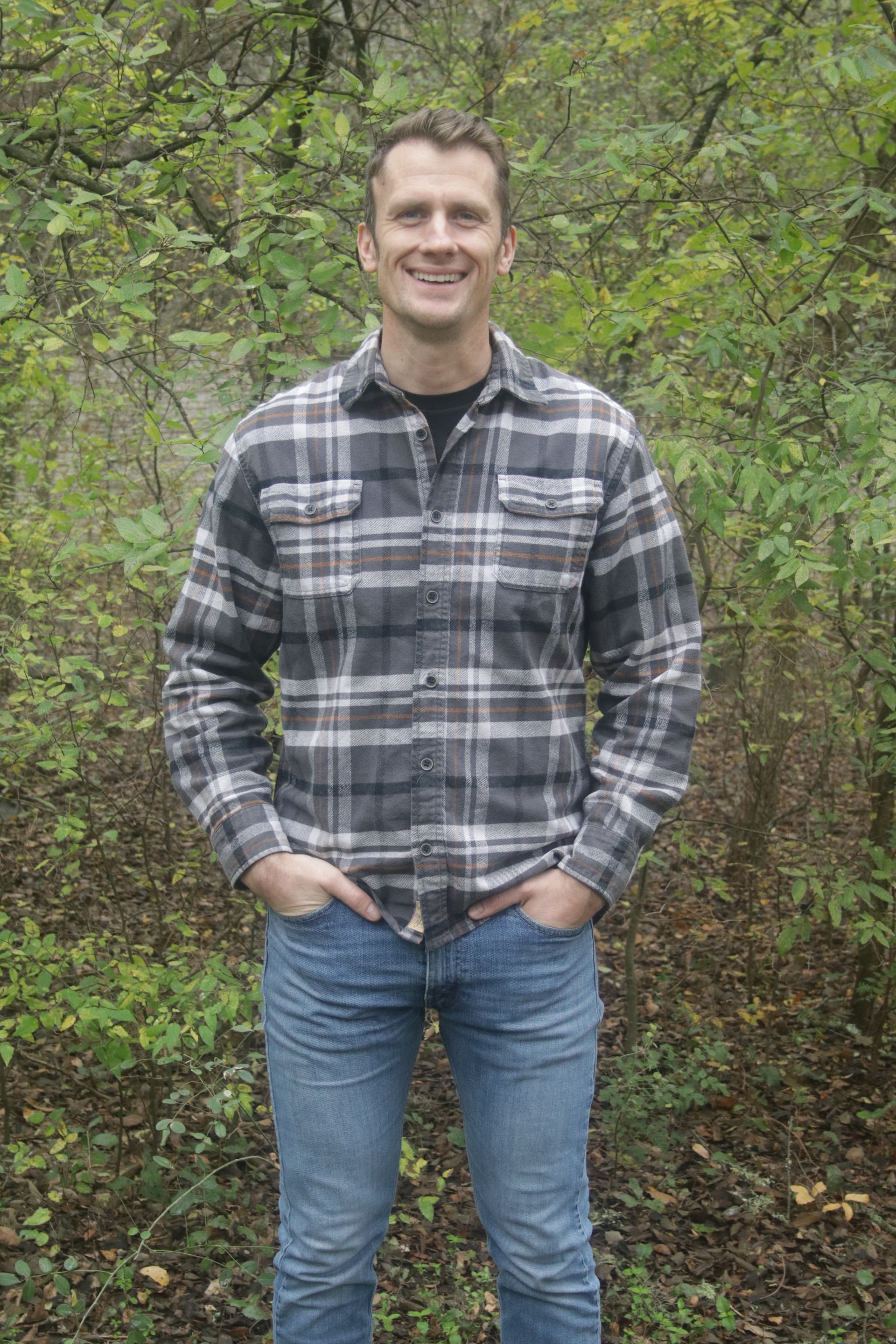 A man in a plaid shirt and jeans is standing in the woods with his hands in his pockets.