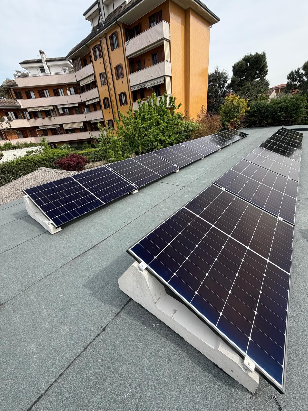 Pannelli solari installati su un tetto verde inclinato accanto a un condominio, con alberi sullo sfondo.