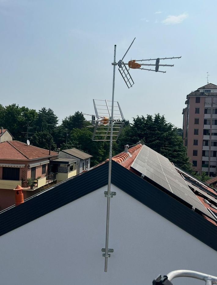 Antenna televisiva sul tetto, sopra i tetti di tegole, con palazzi e alberi sullo sfondo.