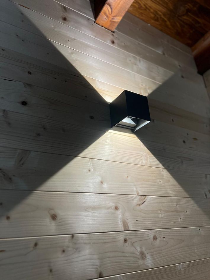Applique nera che proietta una luce triangolare su una parete di assi di legno sotto un soffitto inclinato.