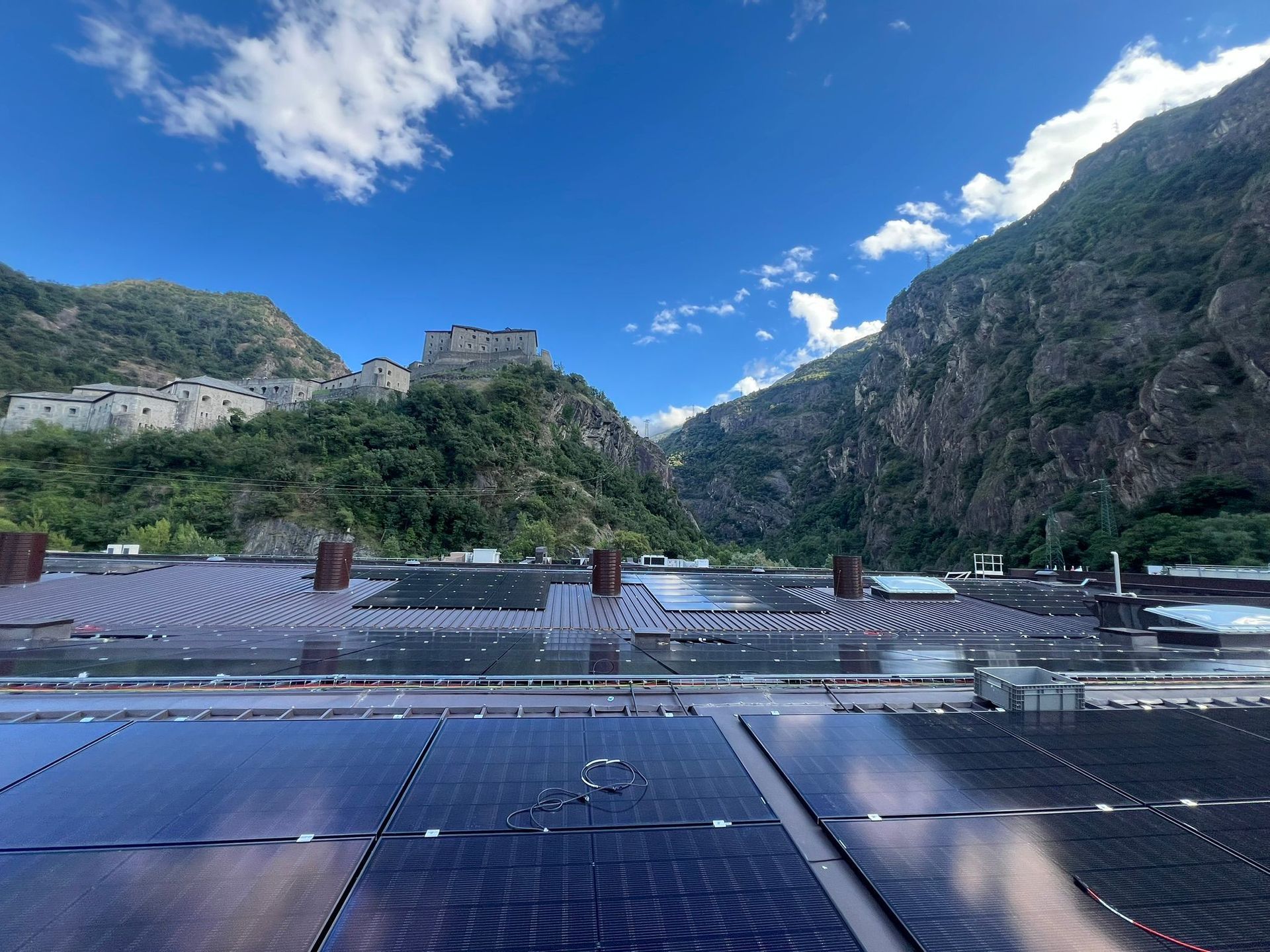 Valle di montagna con edifici in pietra, cielo azzurro e un impianto di pannelli solari sul tetto in primo piano.
