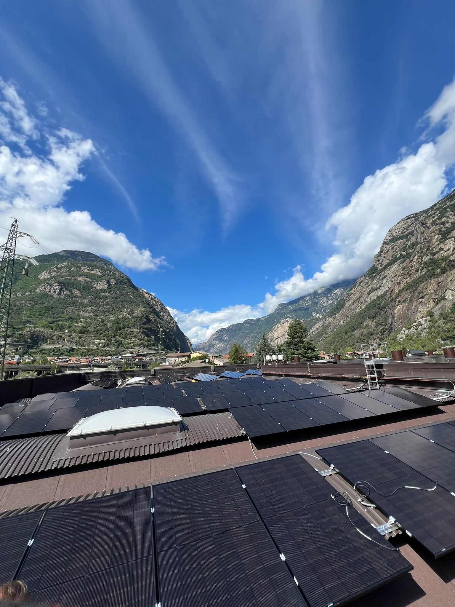 Valle di montagna con pannelli solari in primo piano sotto un cielo azzurro brillante con nuvole leggere.