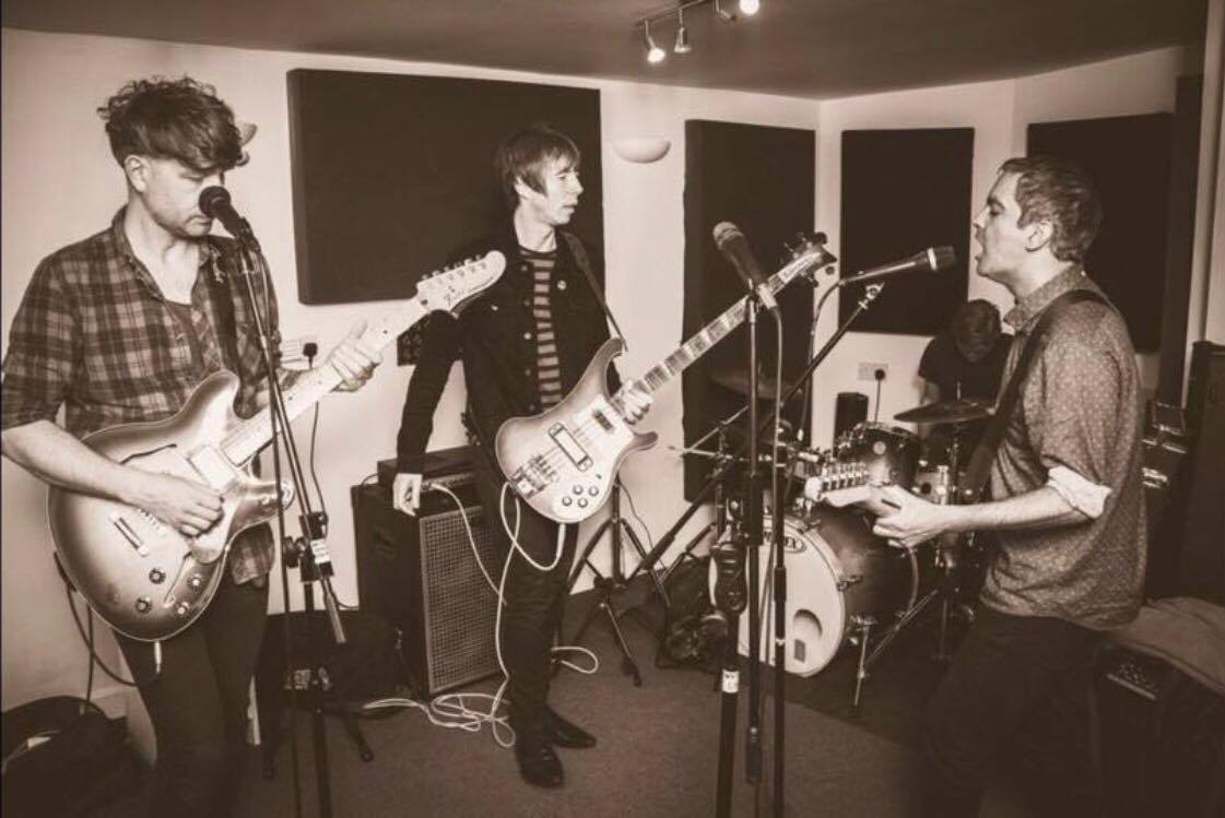 Three men are playing guitars and singing into microphones in a room.