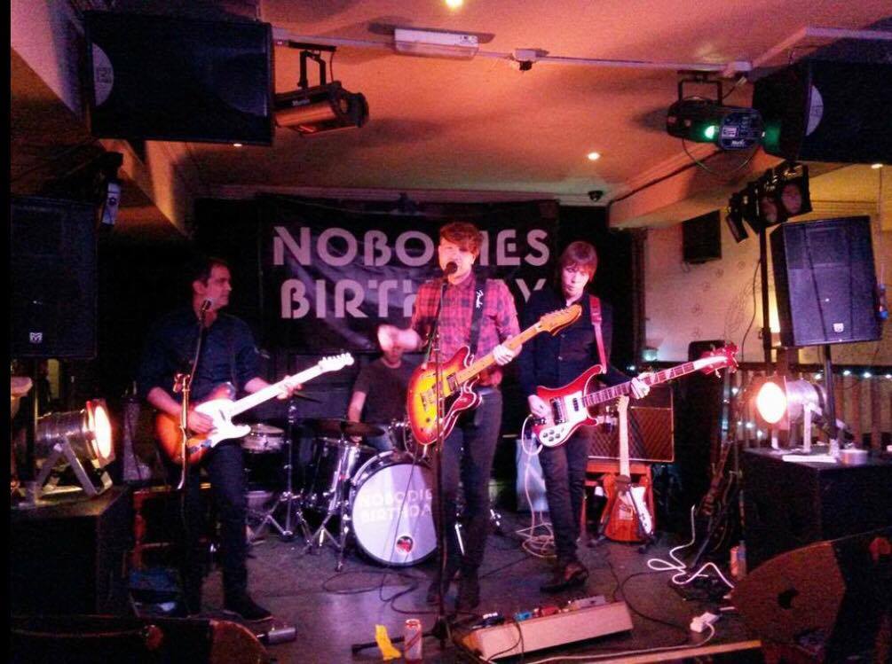 A group of people playing guitars in front of a sign that says nobones birthday