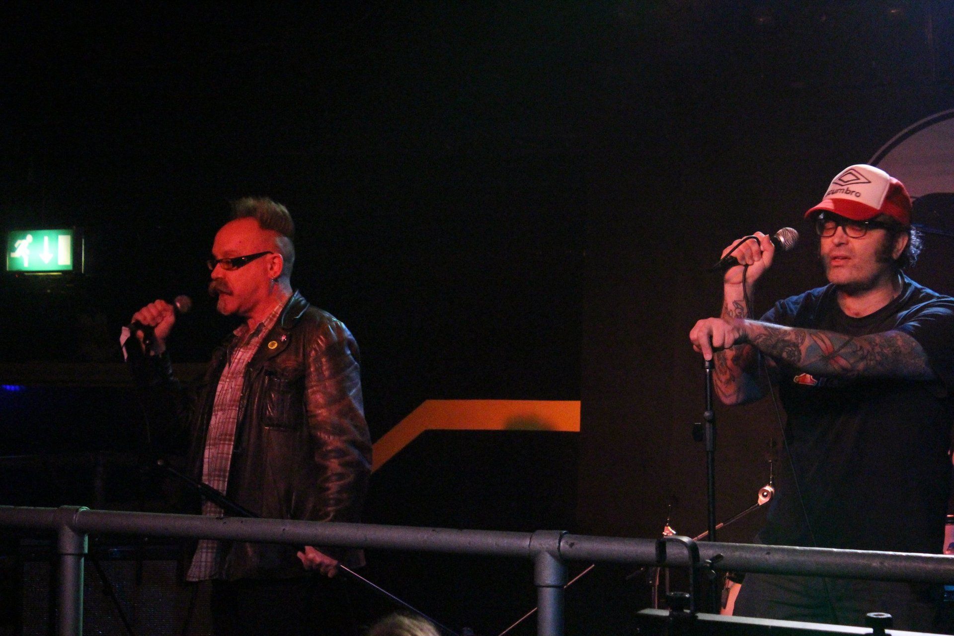 Two men are singing into microphones on a stage in a dark room.