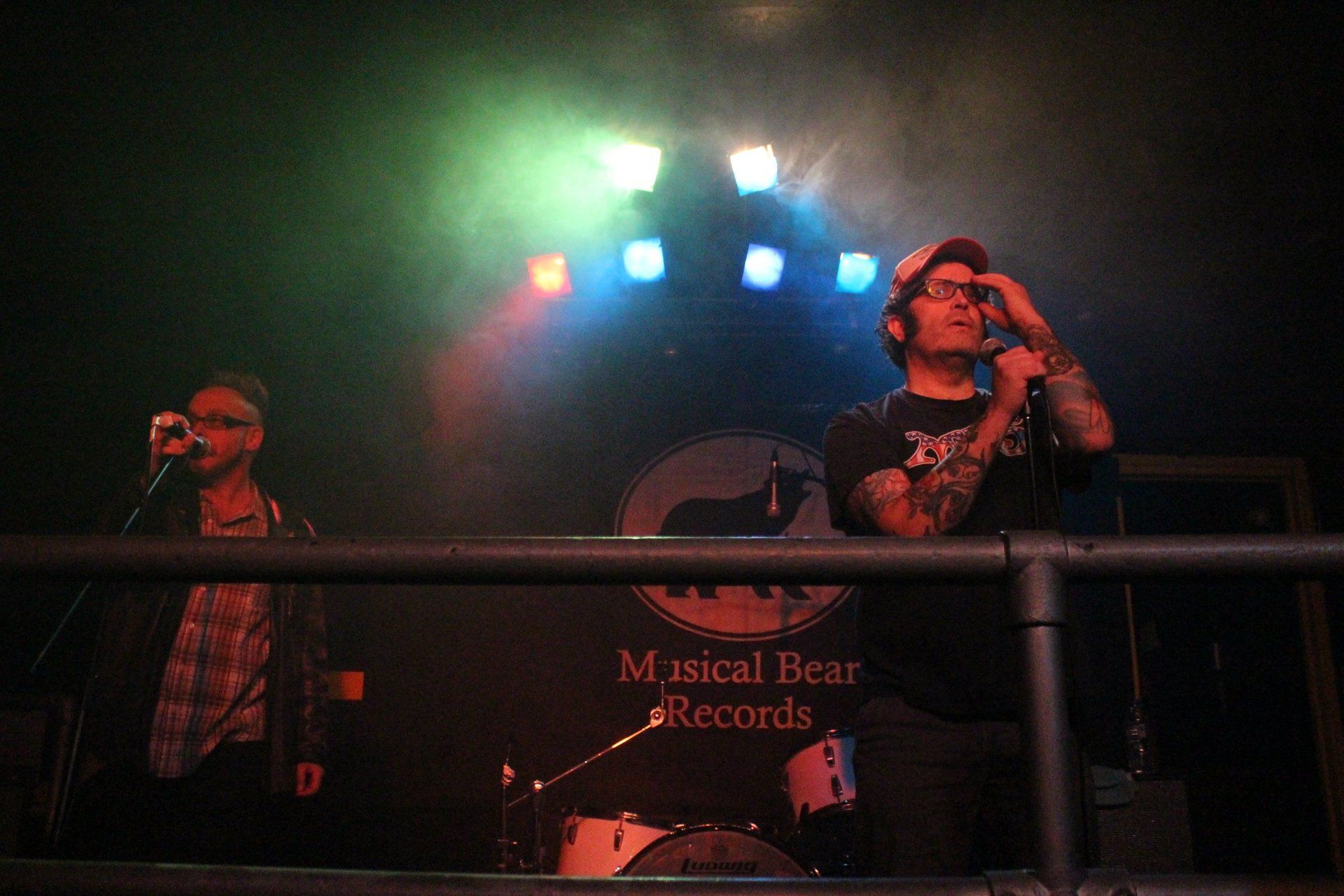 Two men are singing into microphones on a stage in front of a sign that says musical bear records.