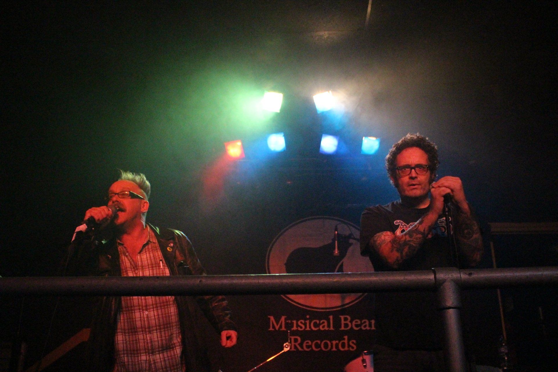 Two men are singing into microphones on a stage in front of a sign that says musical bear records.