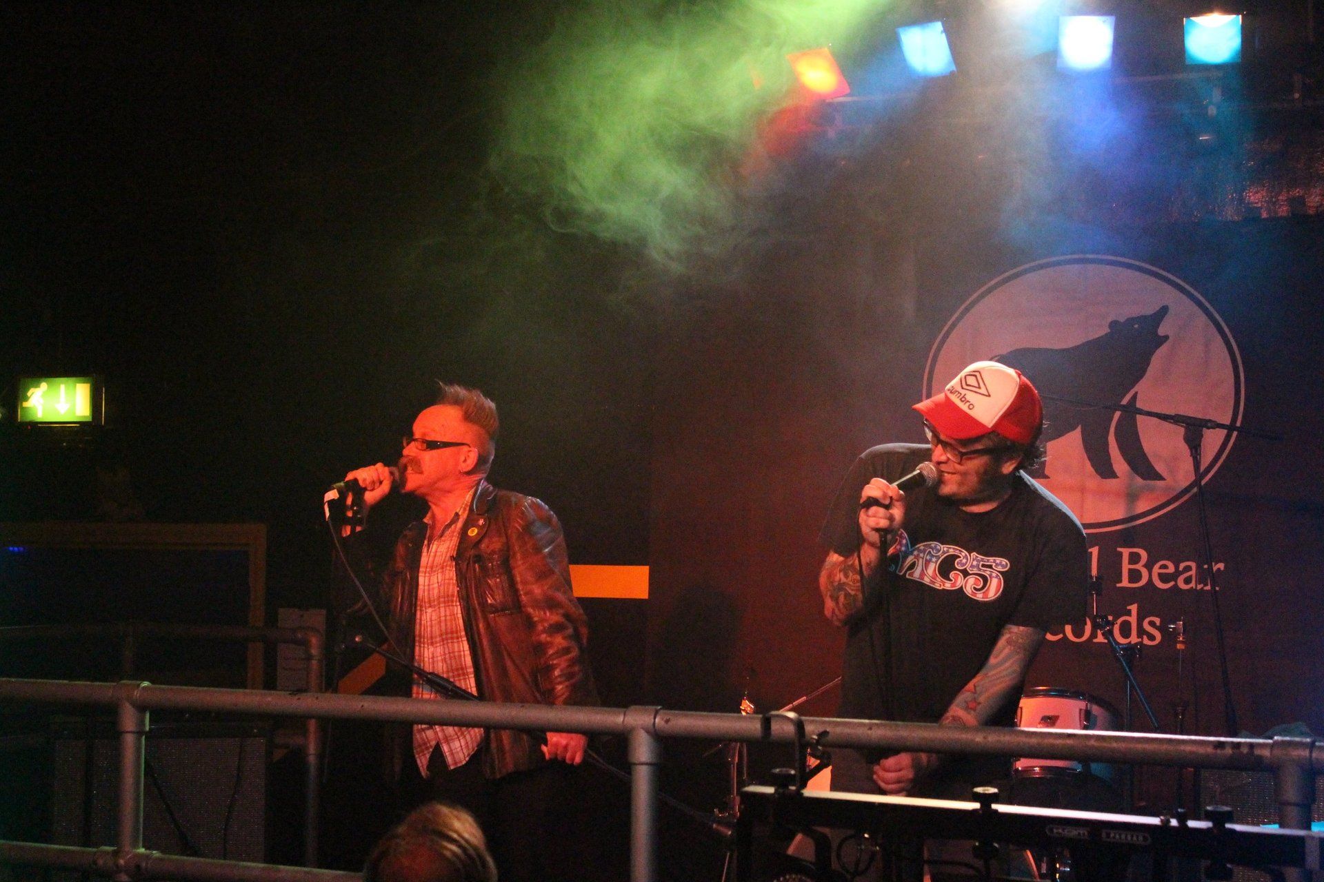 Two men singing into microphones on a stage in front of a sign that says bea