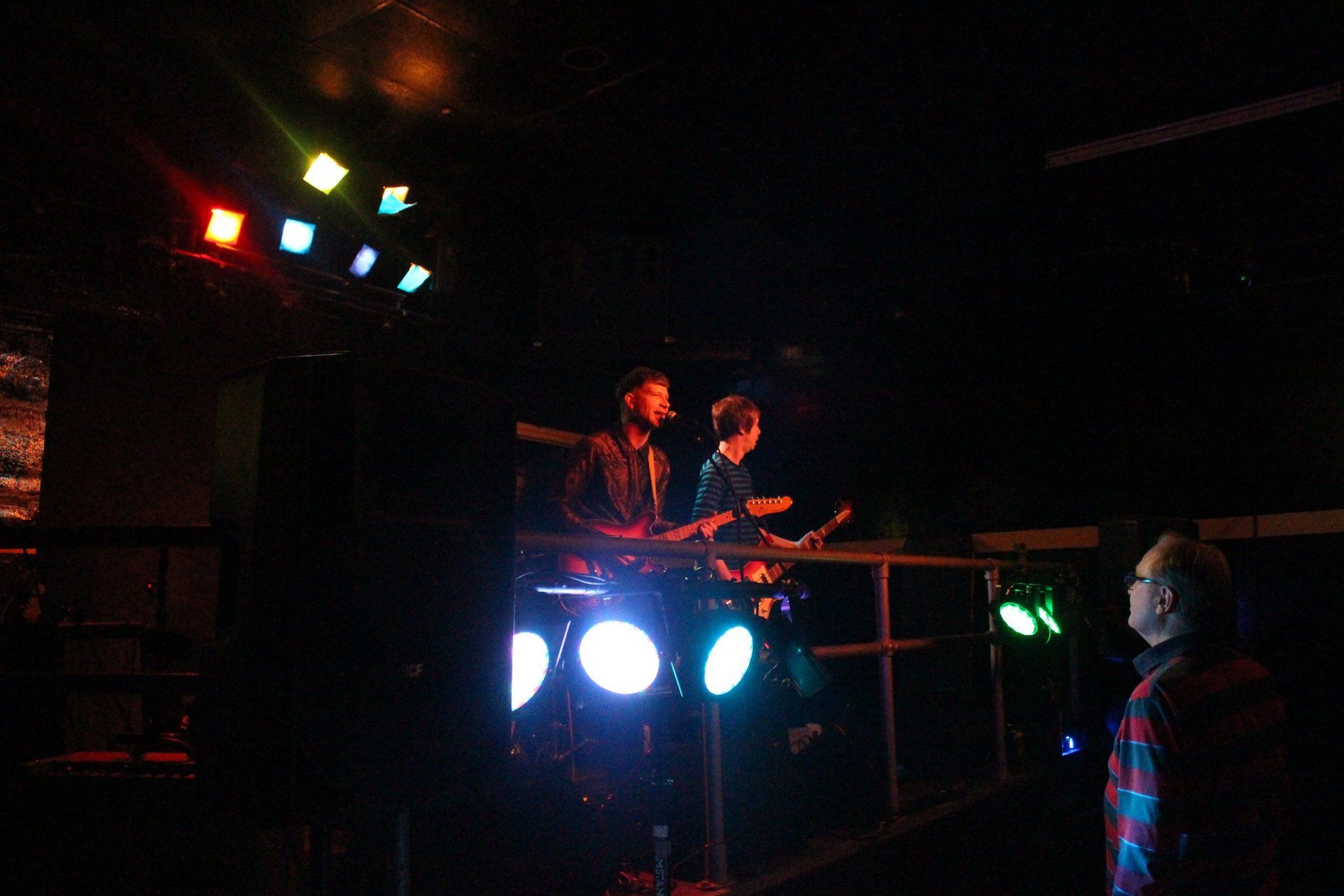 Two men are playing guitars on a stage in a dark room.