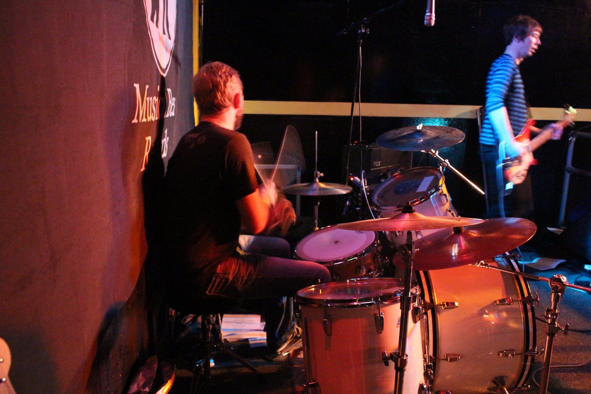 A man playing drums and another man playing guitar on stage