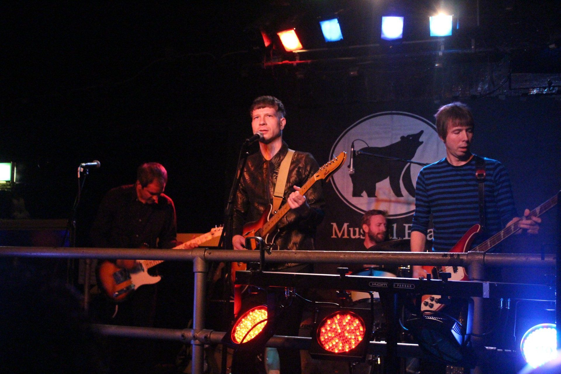 A group of men are playing guitars on a stage in front of a sign that says mu1t8