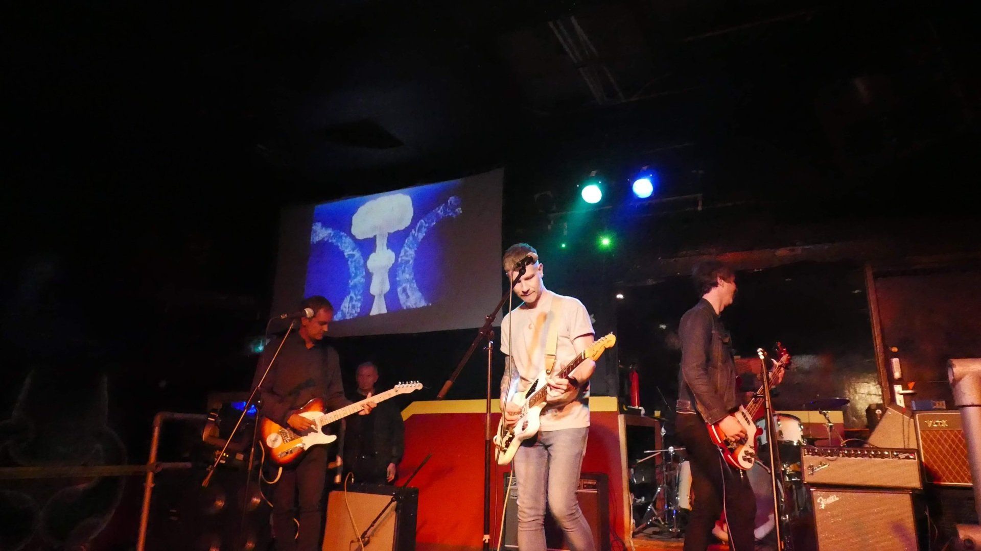 A group of people are playing guitars on a stage in front of a large screen.