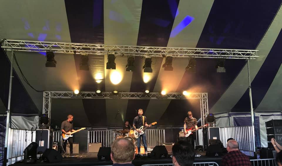A group of people are playing guitars on a stage under a striped tent.