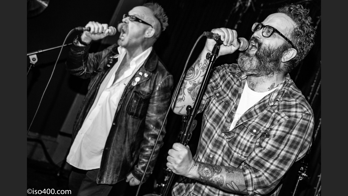 Two men are singing into microphones on a stage in a black and white photo.