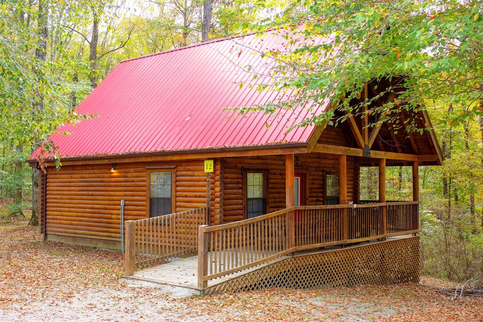Log cabin with a red roof, surrounded by trees. Wooden porch with a ramp.