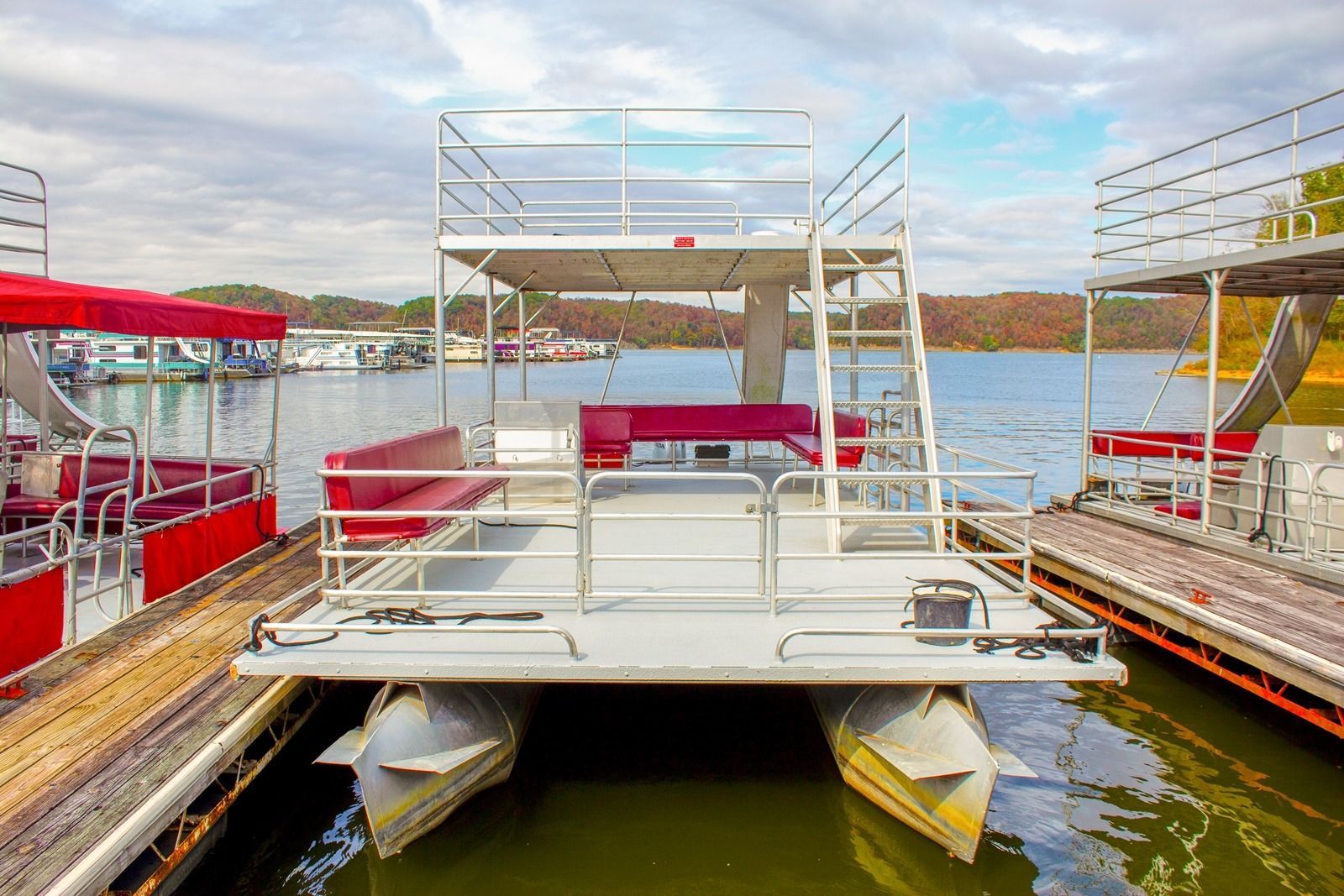 Pontoon boat docked at a marina, with red seating and a second-level deck. Overcast sky, lake background.