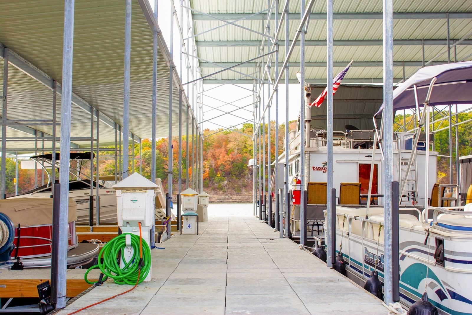 Boats docked in a covered marina, pathway in between. Green hose, American flag, autumn trees in background.