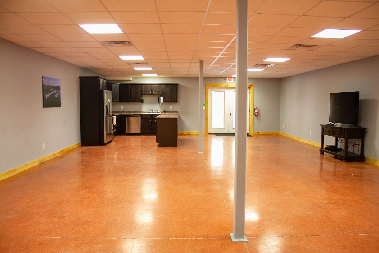 Spacious room with orange floor, kitchen, and TV. Gray walls, overhead lights, and support beams.