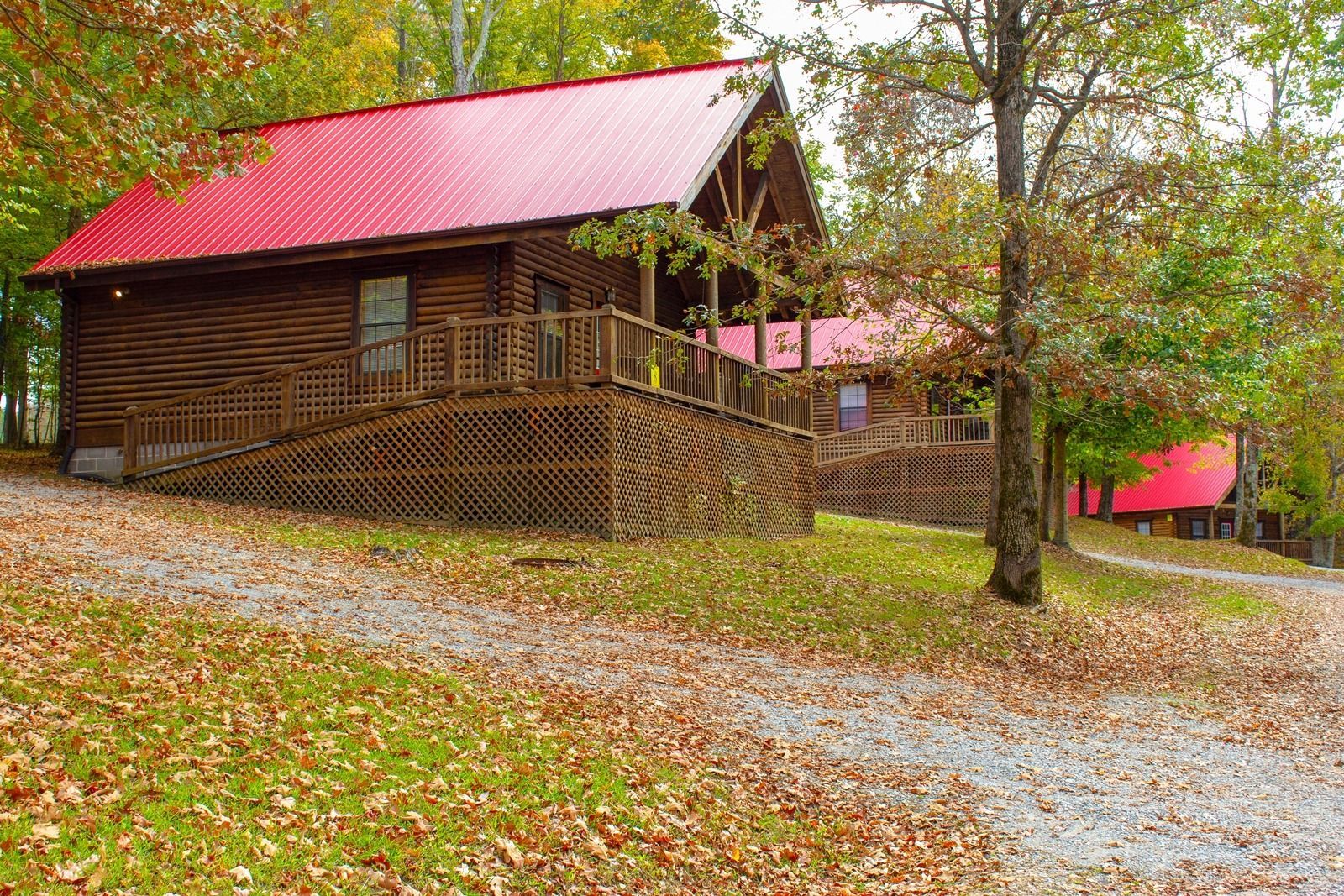 Log cabin with red roof and porch, surrounded by fall foliage and gravel path.