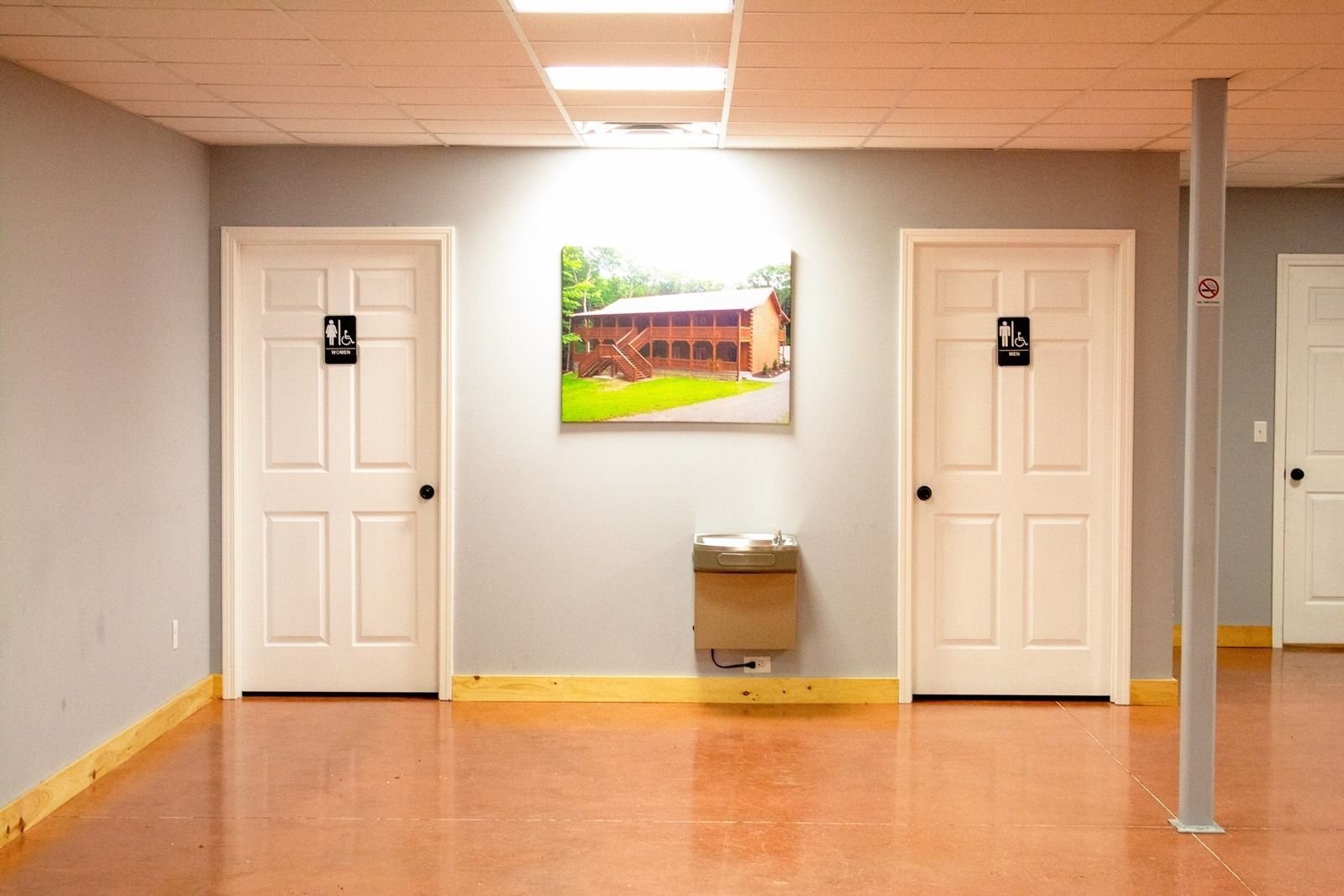 A brightly lit restroom hallway with two white doors, a water fountain, and a photo.