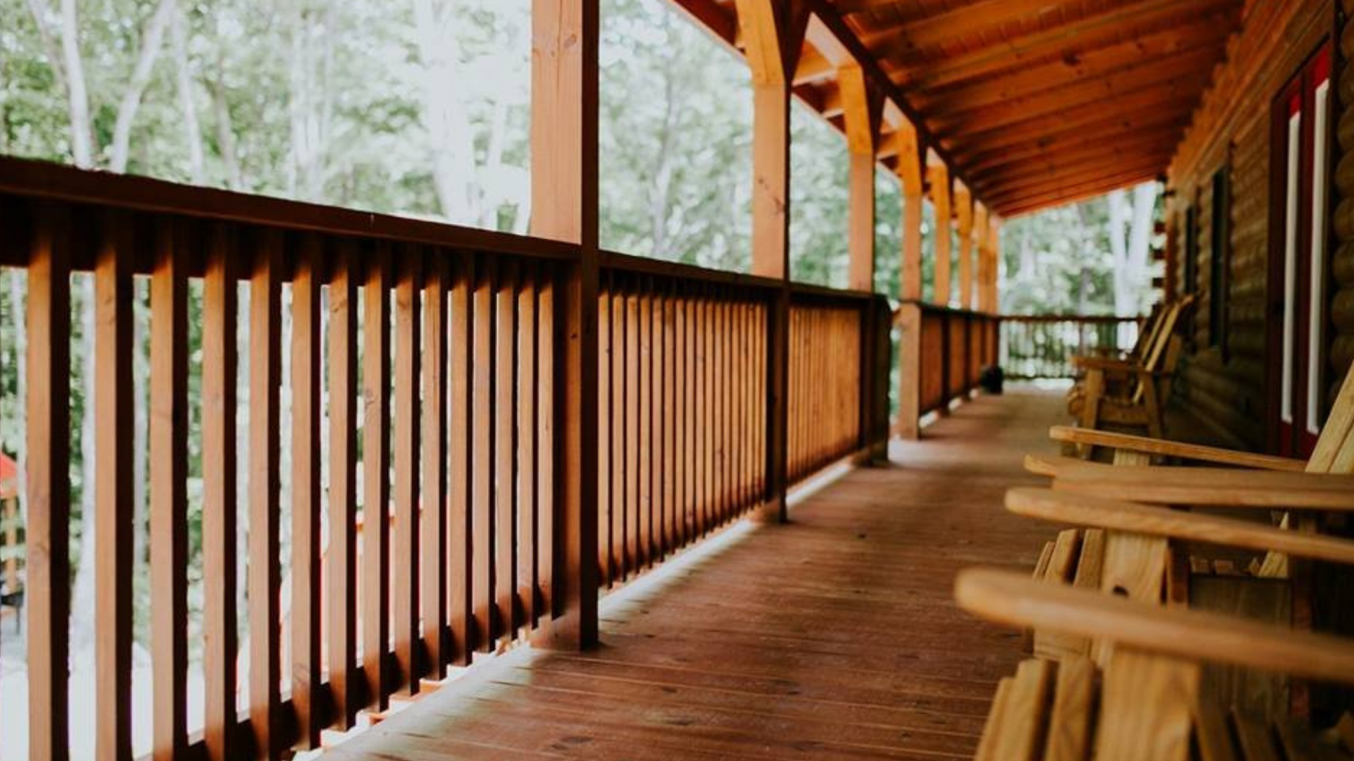 Wooden porch with railing, Adirondack chairs, and log cabin wall; forest background.