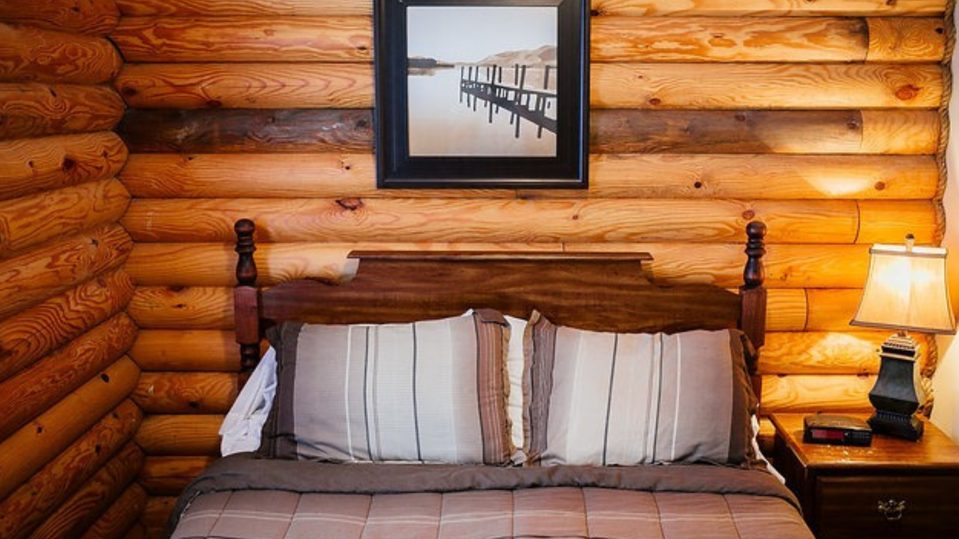 Bedroom with a wooden log wall, bed with pillows, framed picture, and a bedside lamp.